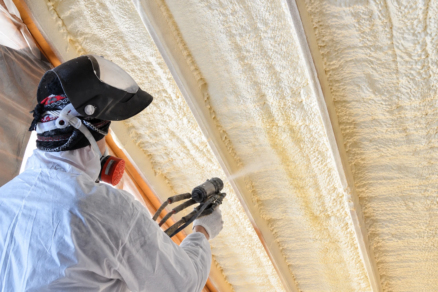 Worker in protective gear applies spray foam to the underside of a roof between exposed rafters.