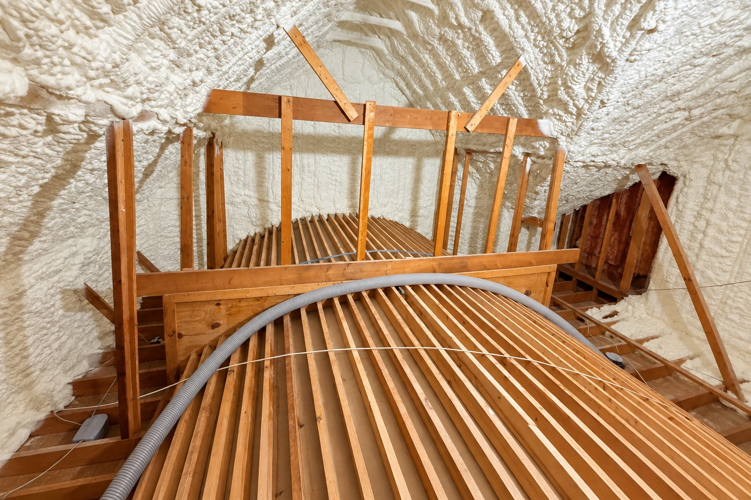 Attic rafters and walls coated in white spray foam, with a flexible hose running across the wooden framing.