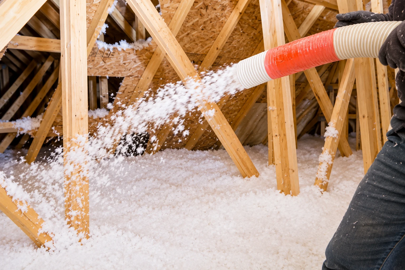 A hose blows loose white insulation into an attic, where it collects across the floor between wooden trusses.
