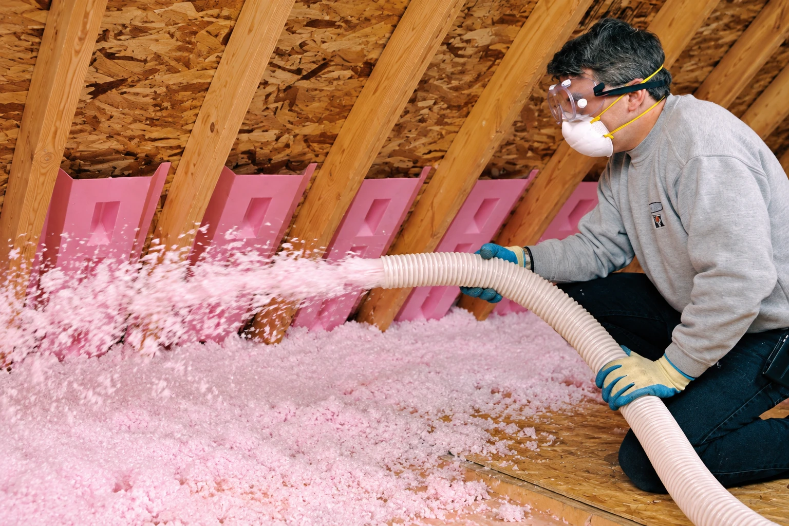 A worker wearing goggles and a respirator blows pink loose-fill insulation into an attic through a flexible hose.