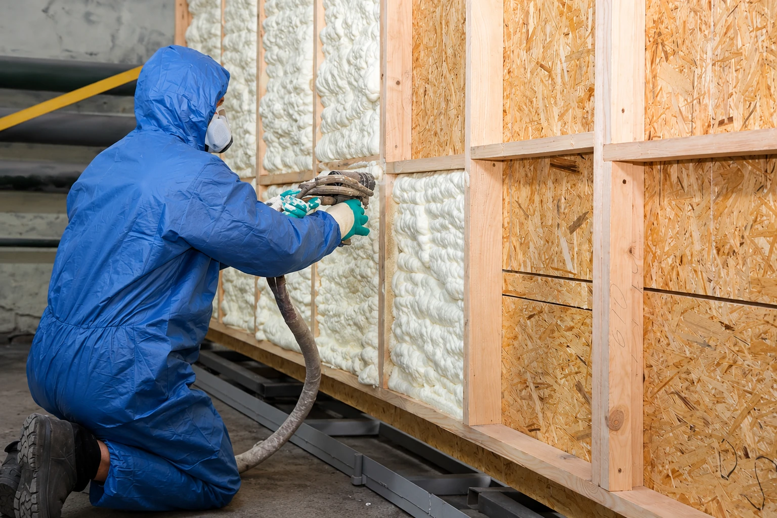 A worker in a blue protective suit sprays foam insulation into the wall cavities of a wood-framed structure.