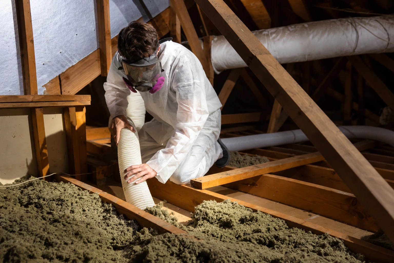 A worker in protective coveralls and a respirator uses a hose to remove loose insulation from an attic.