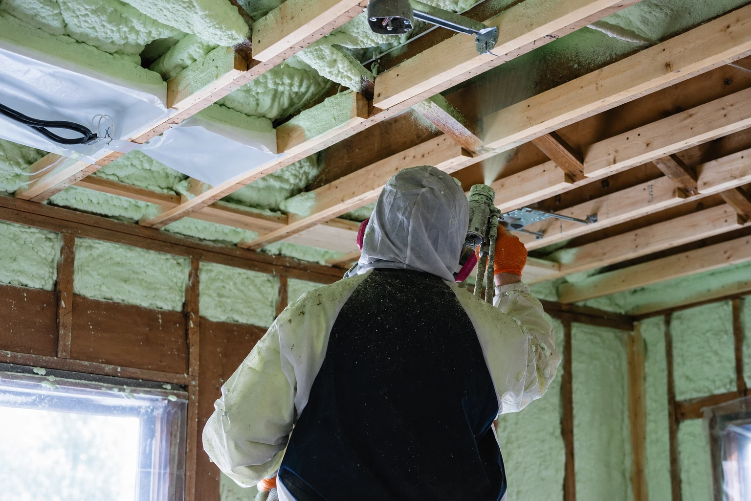 A worker in protective gear sprays pale green foam insulation into exposed ceiling joists.