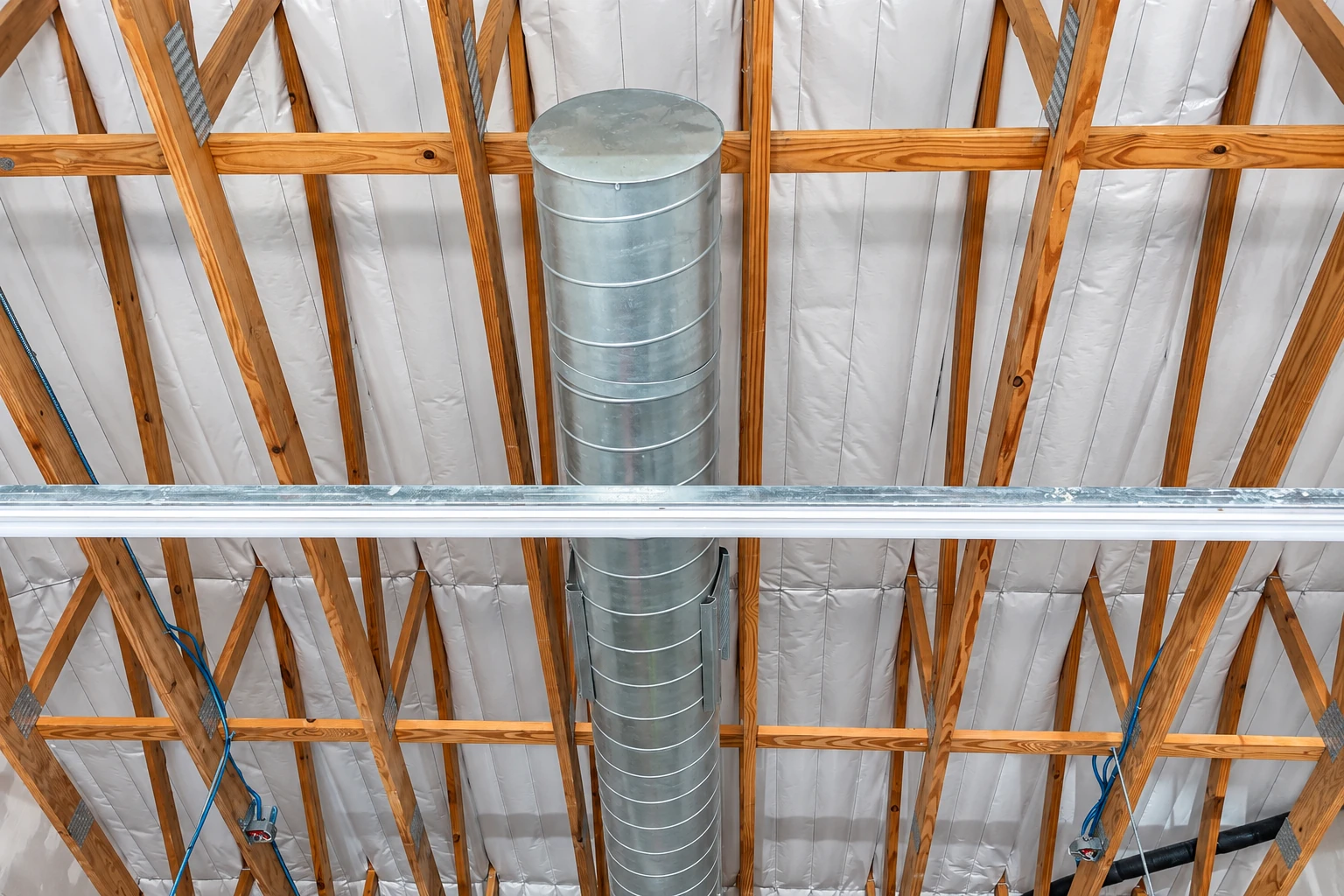 White vapor barrier lines a timber-framed ceiling around a large central metal duct.